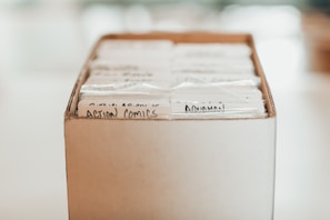 A cardboard box filled with comic books that are neatly organized and labeled with handwritten tags. The comics are protected in plastic sleeves, and the labels include titles like 'Action Comics' written in black ink.