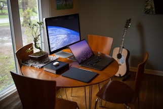 A cozy workspace is set up with a wooden table and chairs near a large window. On the table are a large computer monitor displaying an image of Earth from space, a laptop showing a city skyline at dusk, and a few tech accessories like a wireless mouse and notebook. A small potted plant adds a touch of nature, and an acoustic guitar leans against the wall in the corner.