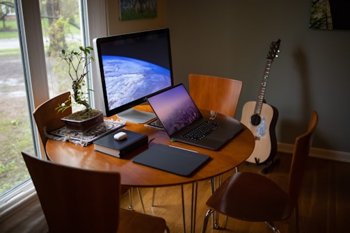 A cozy workspace is set up with a wooden table and chairs near a large window. On the table are a large computer monitor displaying an image of Earth from space, a laptop showing a city skyline at dusk, and a few tech accessories like a wireless mouse and notebook. A small potted plant adds a touch of nature, and an acoustic guitar leans against the wall in the corner.