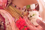 Intricate traditional jewelry adorns the neck, featuring a gold necklace with red and green gemstones. A beaded necklace, also in red, white, and green, is layered above. The image includes vibrant red and white flowers, likely a part of ceremonial attire, along with detailed embroidery on fabric.