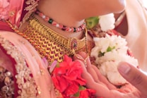 Intricate traditional jewelry adorns the neck, featuring a gold necklace with red and green gemstones. A beaded necklace, also in red, white, and green, is layered above. The image includes vibrant red and white flowers, likely a part of ceremonial attire, along with detailed embroidery on fabric.