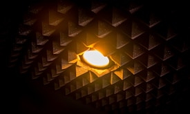 A warm, glowing light bulb set against a dark, textured wall with a pattern of triangular protrusions. The illumination casts a soft orange-yellow glow on the surrounding surface, creating a cozy ambiance.