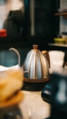 A stainless steel kettle with a wooden handle and base is placed on a countertop. The spout is long and curved, suitable for precise pouring. In the background, there are blurred shapes of kitchen utensils and storage containers, suggesting a kitchen or café setting.
