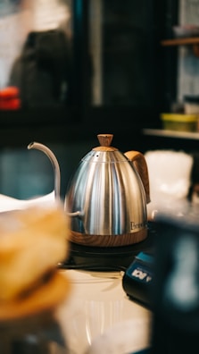 A stainless steel kettle with a wooden handle and base is placed on a countertop. The spout is long and curved, suitable for precise pouring. In the background, there are blurred shapes of kitchen utensils and storage containers, suggesting a kitchen or café setting.