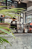 A cozy cafe interior with a menu board displaying various coffee and tea options. A barista stands behind the counter, surrounded by coffee brewing equipment, cups, and plants. Bright natural light streams in through the window, highlighting the rustic decor and indoor greenery.