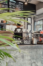 A cozy cafe interior with a menu board displaying various coffee and tea options. A barista stands behind the counter, surrounded by coffee brewing equipment, cups, and plants. Bright natural light streams in through the window, highlighting the rustic decor and indoor greenery.