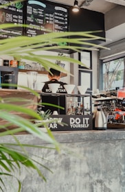 A cozy cafe interior with a menu board displaying various coffee and tea options. A barista stands behind the counter, surrounded by coffee brewing equipment, cups, and plants. Bright natural light streams in through the window, highlighting the rustic decor and indoor greenery.