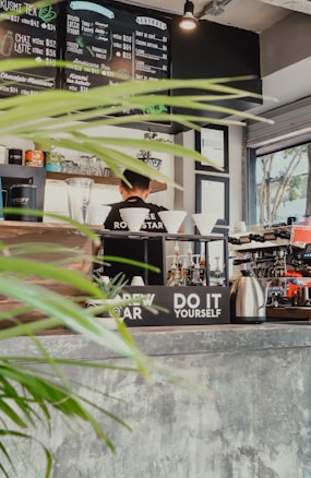 A cozy cafe interior with a menu board displaying various coffee and tea options. A barista stands behind the counter, surrounded by coffee brewing equipment, cups, and plants. Bright natural light streams in through the window, highlighting the rustic decor and indoor greenery.