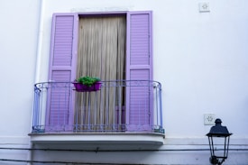 A small balcony with ornate railings painted in a lavender color. The balcony features a matching lavender window with shutters and a vertical bamboo curtain. A purple planter with green foliage sits on the ledge. The facade is white with minimal decoration, and a black lantern is mounted on the wall.