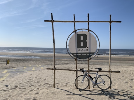 A seaside scene featuring a bicycle resting against a wooden structure with a circular sign displaying the word 'B.E.A.C.H.'. The sandy beach leads to a calm ocean under a clear blue sky.