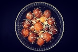 A glass bowl filled with round, brown, syrupy sweets, sprinkled with slivers of nuts, likely served as a dessert.