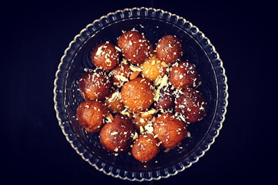 A glass bowl filled with round, brown, syrupy sweets, sprinkled with slivers of nuts, likely served as a dessert.