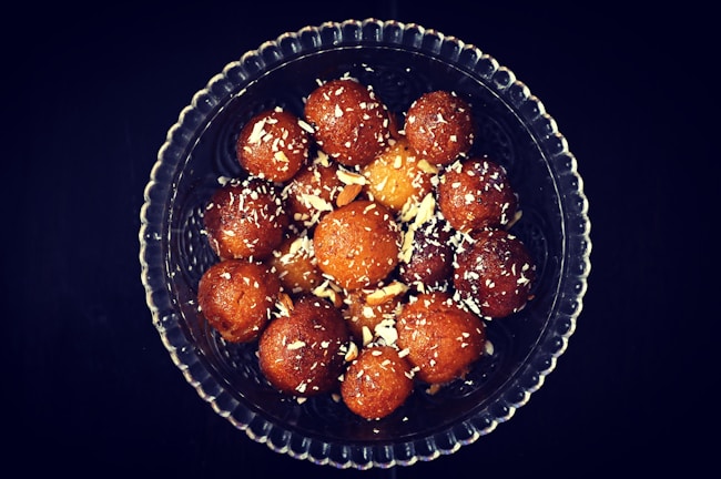 A glass bowl filled with round, brown, syrupy sweets, sprinkled with slivers of nuts, likely served as a dessert.