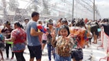 A large group of people engaging in a water festival, covered in foam. Many are smiling and having fun, with one person in the foreground making a peace sign. The scene is lively, with colorful water guns and vibrant clothing. Decorations hang overhead, adding to the festive atmosphere.
