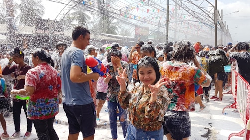 A large group of people engaging in a water festival, covered in foam. Many are smiling and having fun, with one person in the foreground making a peace sign. The scene is lively, with colorful water guns and vibrant clothing. Decorations hang overhead, adding to the festive atmosphere.