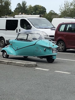 A vintage three-wheeled car with a turquoise body is parked in a lot near other vehicles, including a white van and a red car. The car has a compact design with a clear dome on top. Trees and other greenery are visible in the background.