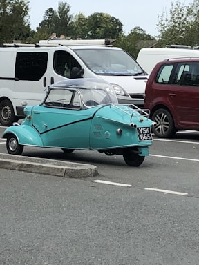 A vintage three-wheeled car with a turquoise body is parked in a lot near other vehicles, including a white van and a red car. The car has a compact design with a clear dome on top. Trees and other greenery are visible in the background.