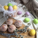 Pastel-colored eggs are nestled in a wicker basket filled with white shredded paper, and a few are scattered on a cloth nearby. Four glazed donuts topped with colorful sprinkles are positioned on a wooden board, alongside soft, fuzzy pussy willow branches.