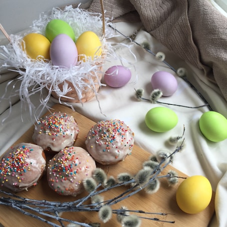 Pastel-colored eggs are nestled in a wicker basket filled with white shredded paper, and a few are scattered on a cloth nearby. Four glazed donuts topped with colorful sprinkles are positioned on a wooden board, alongside soft, fuzzy pussy willow branches.
