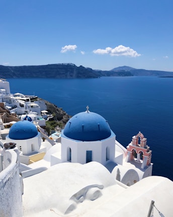 A picturesque seaside view of white buildings with blue domes and details, perched on a cliff overlooking a deep blue sea. In the background, distant mountains create a serene and majestic backdrop against a clear blue sky with a few fluffy clouds.