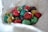 A collection of colorful, speckled eggs nestled in a soft, white, textured cloth. The eggs vary in color, featuring shades of red, yellow, green, blue, and purple, each with black speckles that add to their decorative appearance.