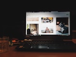 A laptop displaying a website with various images including a woman sitting indoors, a moving race car, a cabin in a mountainous area, and a dog. The device is placed on a wooden table with a glass of water and a notebook nearby, in a dimly lit environment.