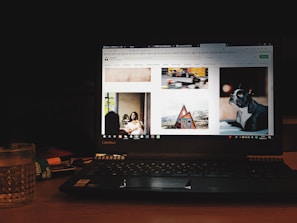 A laptop displaying a website with various images including a woman sitting indoors, a moving race car, a cabin in a mountainous area, and a dog. The device is placed on a wooden table with a glass of water and a notebook nearby, in a dimly lit environment.