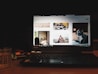 A laptop displaying a website with various images including a woman sitting indoors, a moving race car, a cabin in a mountainous area, and a dog. The device is placed on a wooden table with a glass of water and a notebook nearby, in a dimly lit environment.