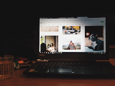 A laptop displaying a website with various images including a woman sitting indoors, a moving race car, a cabin in a mountainous area, and a dog. The device is placed on a wooden table with a glass of water and a notebook nearby, in a dimly lit environment.