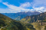 A breathtaking mountain landscape with towering peaks partially covered in snow under a bright blue sky with wispy clouds. A narrow winding road cuts through lush green valleys and pine forests, offering a sense of scale and adventure.