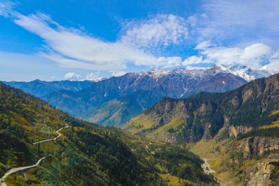 A breathtaking mountain landscape with towering peaks partially covered in snow under a bright blue sky with wispy clouds. A narrow winding road cuts through lush green valleys and pine forests, offering a sense of scale and adventure.
