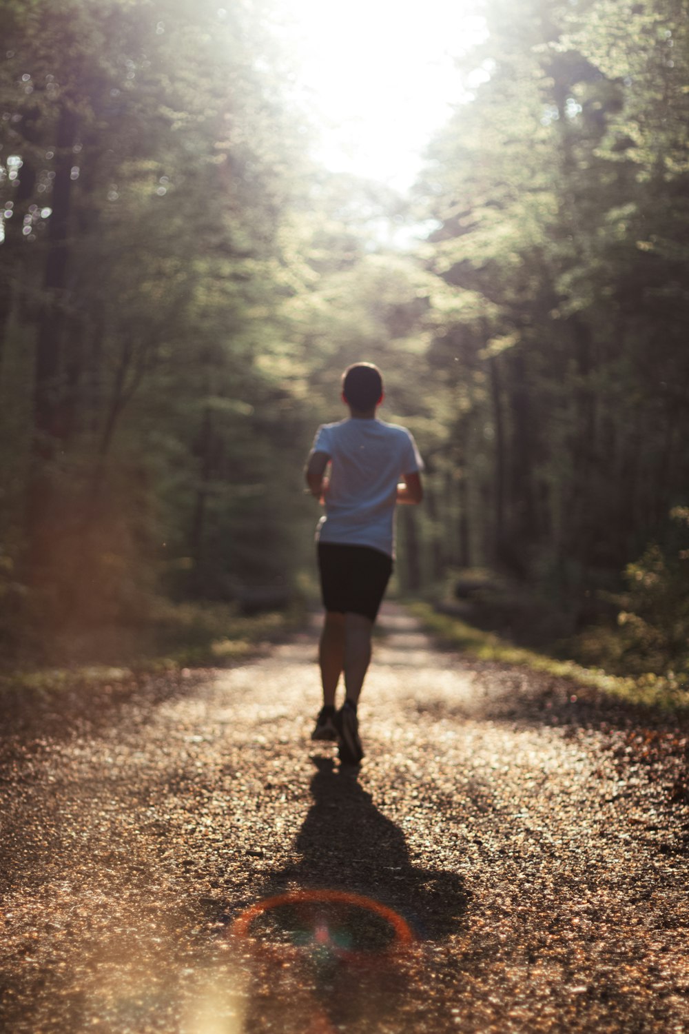 man in white shirt and black shorts walking on dirt road between trees during daytime