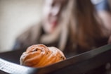 A close-up view of a freshly baked pastry with a flaky texture and a dusting of powdered sugar. The pastry is in the foreground, appearing warm and inviting. The background is blurred, possibly indicating a setting in a cafe or kitchen.
