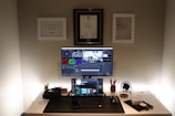 A home office desk setup featuring a large monitor and a laptop displaying editing software. The workspace is organized, with a mouse, keyboard, headphones, and various office supplies. Certificates and awards are mounted on the wall above the desk.