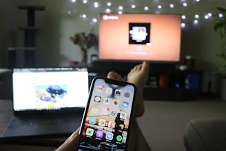 A person is relaxing in a living room with their feet up, holding a smartphone. The screen displays various app icons. A laptop is open on the coffee table in front of them, and a TV is mounted on the wall displaying a media player interface. String lights add a cozy atmosphere.