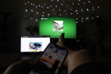 A dimly lit room features a person relaxing with legs stretched out. A television in the background displays a green screen with music playing, while a smartphone is held in the foreground. A laptop is open on a nearby surface displaying a colorful web page. String lights are draped on the wall, adding a cozy ambiance, and potted plants are partially visible.