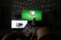 A dimly lit room features a person relaxing with legs stretched out. A television in the background displays a green screen with music playing, while a smartphone is held in the foreground. A laptop is open on a nearby surface displaying a colorful web page. String lights are draped on the wall, adding a cozy ambiance, and potted plants are partially visible.