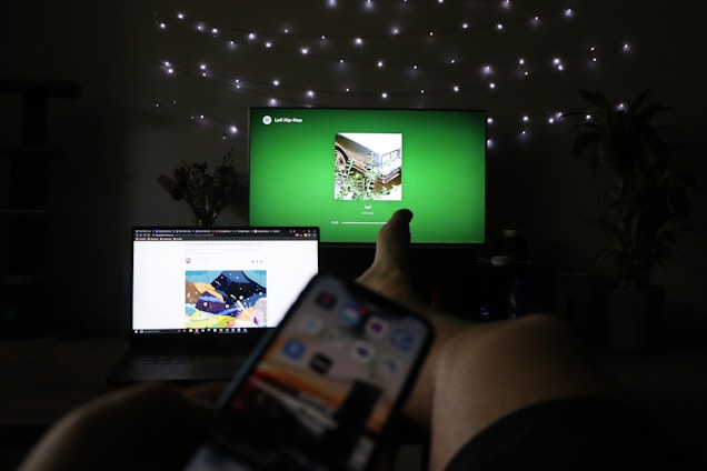 A dimly lit room features a person relaxing with legs stretched out. A television in the background displays a green screen with music playing, while a smartphone is held in the foreground. A laptop is open on a nearby surface displaying a colorful web page. String lights are draped on the wall, adding a cozy ambiance, and potted plants are partially visible.