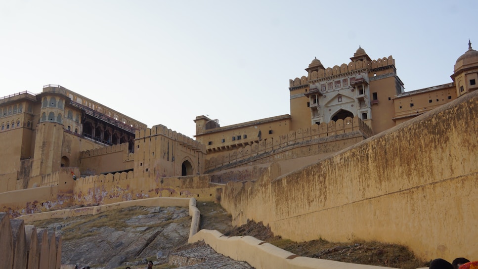A large, historic fortification with a blend of Rajput and Indo-Islamic architectural styles. The structure features multiple levels, arches, and decorative elements, with a slightly weathered appearance. The fortification is situated on a rocky hillside, surrounded by an expansive stone wall.
