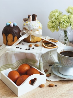 Two decorated pastries are displayed on a cloth with a cup of coffee beside them. One pastry is topped with chocolate glaze and colorful candy eggs, while the other is covered with white icing, sprinkles, and wafer sticks, adorned with a ribbon. Dried orange slices, almonds, and coffee beans are artistically scattered around. A bouquet of light green flowers in a vase adds a touch of elegance to the scene.