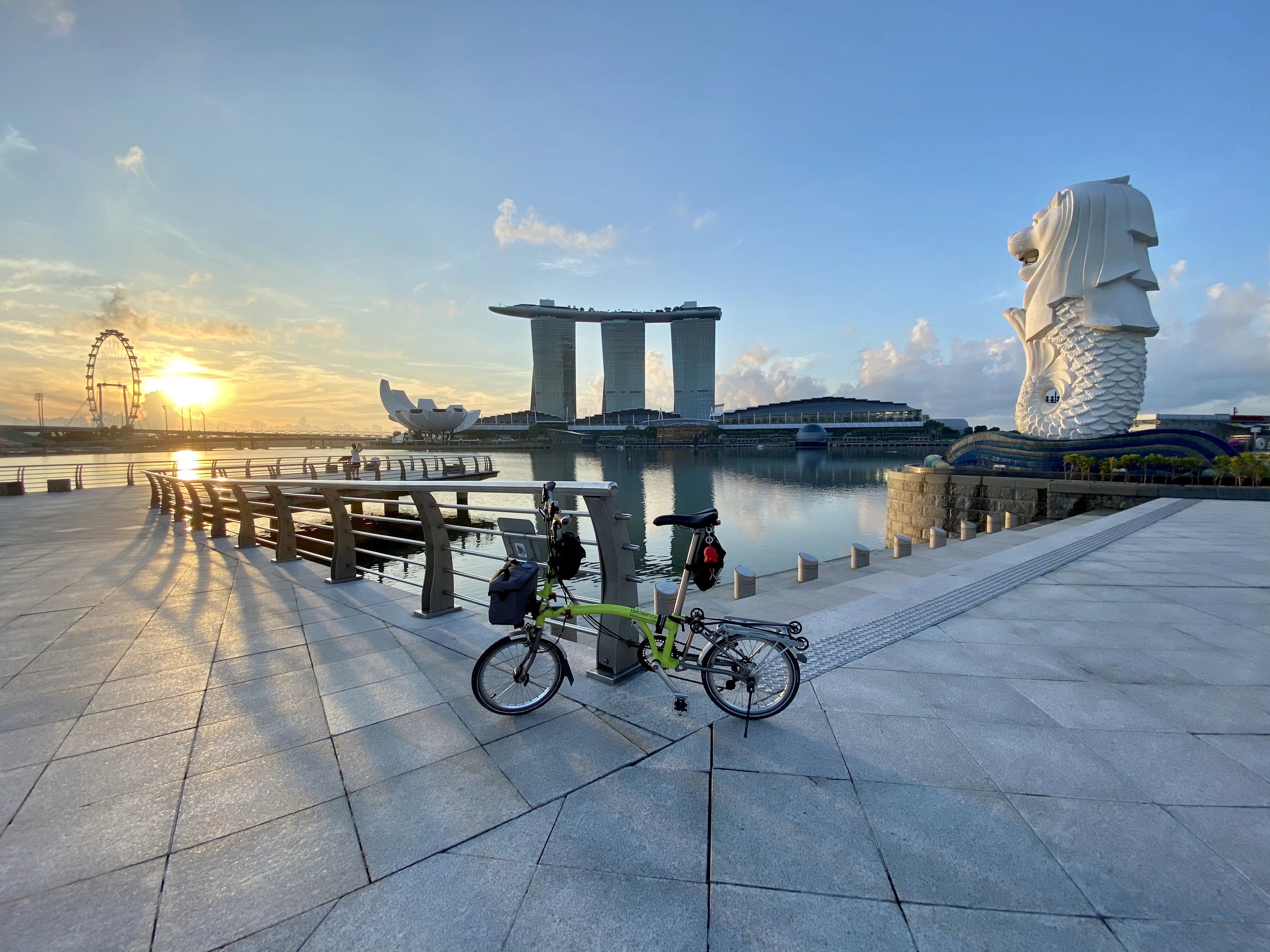 A folding bicycle parked on the waterfront promenade at sunrise, with the Merlion and Marina Bay Sands beyond.