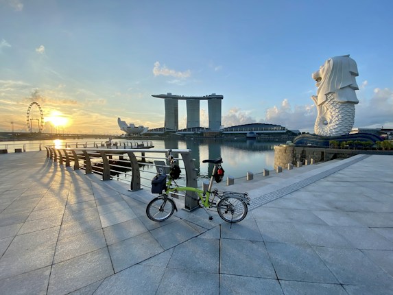 A waterfront scene with the Merlion statue on the right, the Marina Bay Sands hotel with its iconic design in the center, and a ferris wheel to the left. The scene captures the sunrise casting warm colors over the calm water and clear sky, with a bicycle in the foreground on a paved promenade.