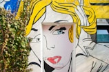 A colorful mural painted on a wall incorporates a door as part of the artwork. The mural features the stylized face of a woman with bright yellow hair and prominent makeup, done in a comic book style. Vines grow on the left side, adding a touch of nature to the scene.