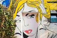 A colorful mural painted on a wall incorporates a door as part of the artwork. The mural features the stylized face of a woman with bright yellow hair and prominent makeup, done in a comic book style. Vines grow on the left side, adding a touch of nature to the scene.