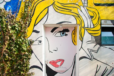 A colorful mural painted on a wall incorporates a door as part of the artwork. The mural features the stylized face of a woman with bright yellow hair and prominent makeup, done in a comic book style. Vines grow on the left side, adding a touch of nature to the scene.