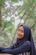A person wearing a dark blue hijab and matching clothing is smiling softly. The background consists of a lush, green, bokeh-filled outdoor setting, suggesting a serene and natural environment.