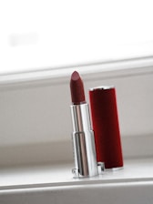 A metallic lipstick tube with a red lipstick inside is placed upright on a white surface. The lipstick casing is partially covered by a red cylindrical cap that stands next to it. The setting is minimalistic with soft lighting coming from a light background, possibly a window.