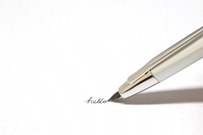 A silver fountain pen is placed on a dotted paper as it writes the word 'hello' in cursive. The pen's sleek metal body contrasts with the simplicity of the paper.