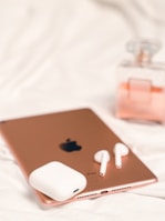 A rose gold tablet with a visible Apple logo is placed on a white surface. Next to the tablet, there are white wireless earbuds and their charging case. In the background, a partially visible glass perfume bottle with a pink hue adds a touch of elegance.