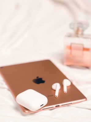 A rose gold tablet with a visible Apple logo is placed on a white surface. Next to the tablet, there are white wireless earbuds and their charging case. In the background, a partially visible glass perfume bottle with a pink hue adds a touch of elegance.